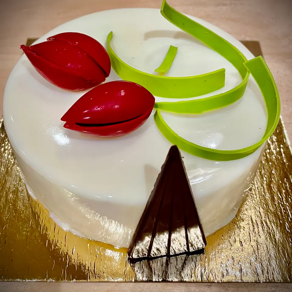 A round cake with a glossy white mirror glaze, decorated with two red tulip-shaped chocolates, green chocolate curls, and a dark chocolate triangle on a gold base.