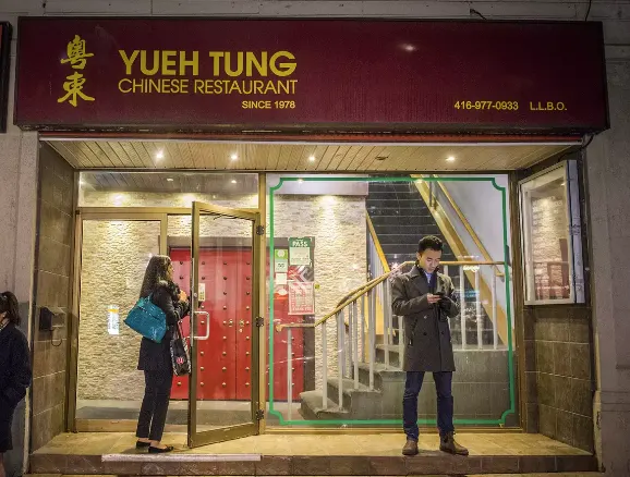 Exterior storefront of Yueh Tung Chinese Restaurant at night, featuring a dark red sign with gold lettering, a glass entrance, and patrons standing outside.