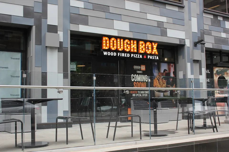 Exterior view of Dough Box Wood Fired Pizza & Pasta in Toronto, featuring a modern grey tiled facade, a bright orange marquee sign, and an outdoor patio with black tables and chairs behind a glass railing.