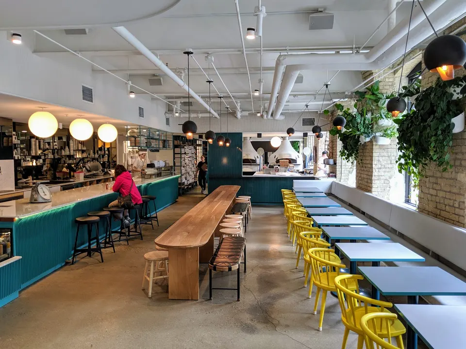 Spacious interior of General Assembly Pizza featuring a long wooden communal table, a row of small tables with bright yellow chairs, a teal service counter with bar stools, and hanging plants.