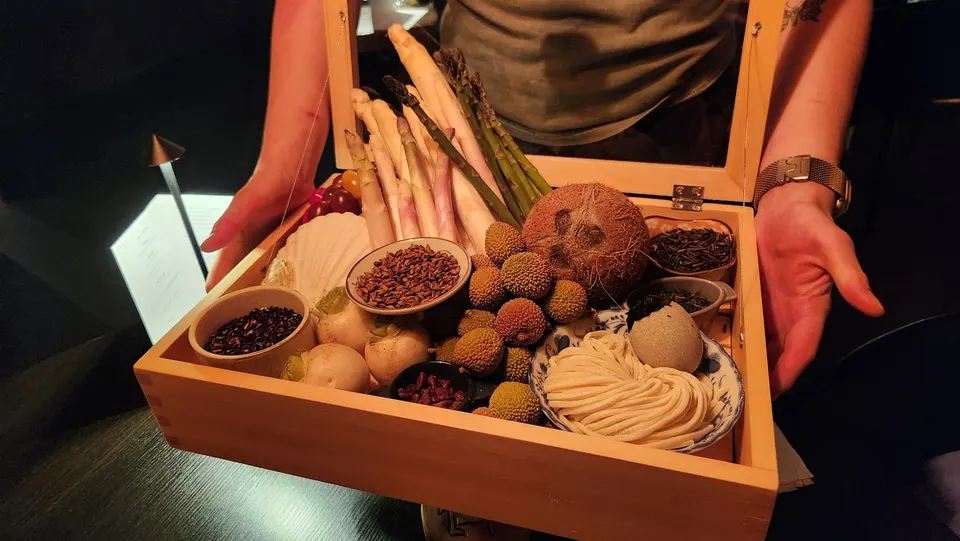A wooden display box held by a server, filled with raw ingredients including white and green asparagus, a whole coconut, lychees, fresh noodles, small bowls of grains and tea leaves, and root vegetables.