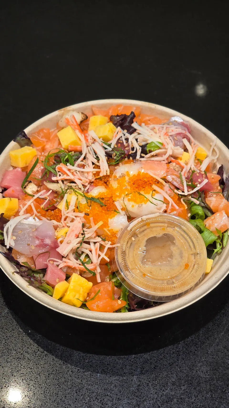 A colorful poke bowl filled with salmon, tuna, crab stick, tamago egg cubes, tobiko, and greens, served with a side of sesame dressing in a plastic cup.
