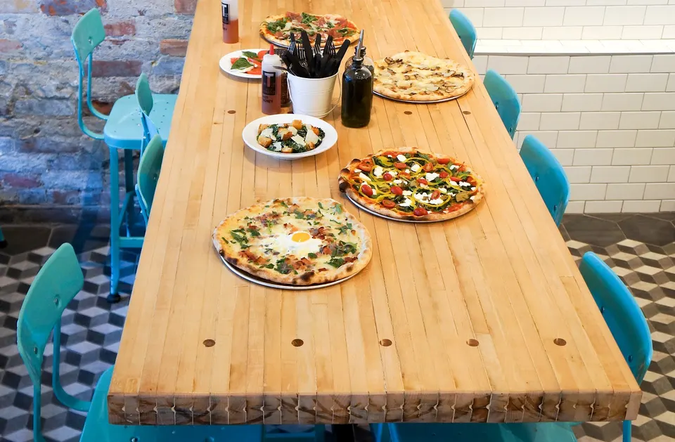 A long wooden table set with several pizzas, a salad, and condiments, surrounded by bright turquoise chairs in a modern pizzeria.