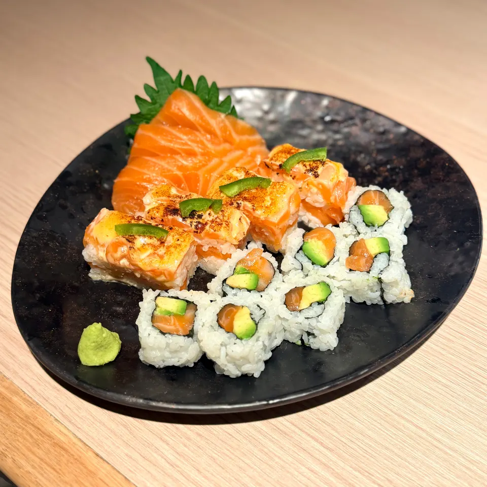 A black plate featuring a selection of sushi, including salmon sashimi on a green leaf, seared salmon rolls topped with green pepper slices, and salmon avocado maki rolls, served with a dollop of wasabi.