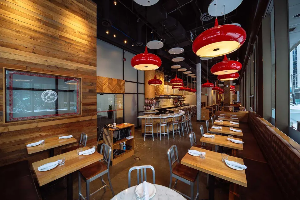 Modern restaurant interior featuring warm wood-paneled walls, sleek wooden tables set with white plates and glasses, metal chairs, and a row of large, glossy red pendant lights hanging over the dining area.