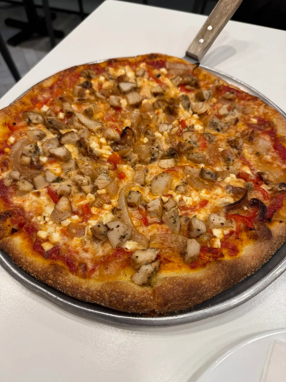 A freshly baked thin-crust pizza topped with grilled chicken, onions, red peppers, and melted cheese, served on a metal tray with a wooden-handled server.