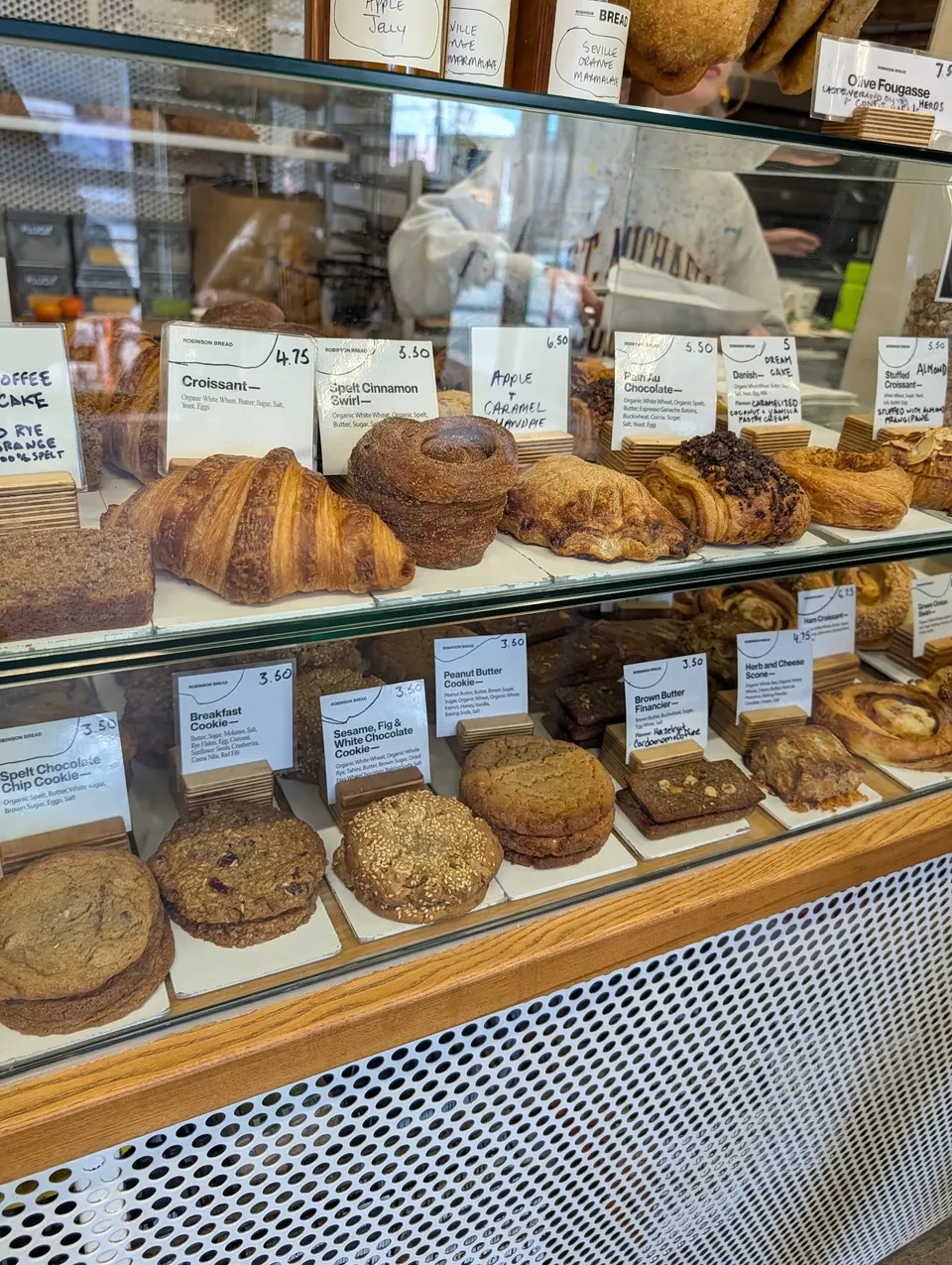 A glass display case at Robinson Bread filled with freshly baked pastries, including croissants, cinnamon swirls, and various cookies, each labeled with small price tags.