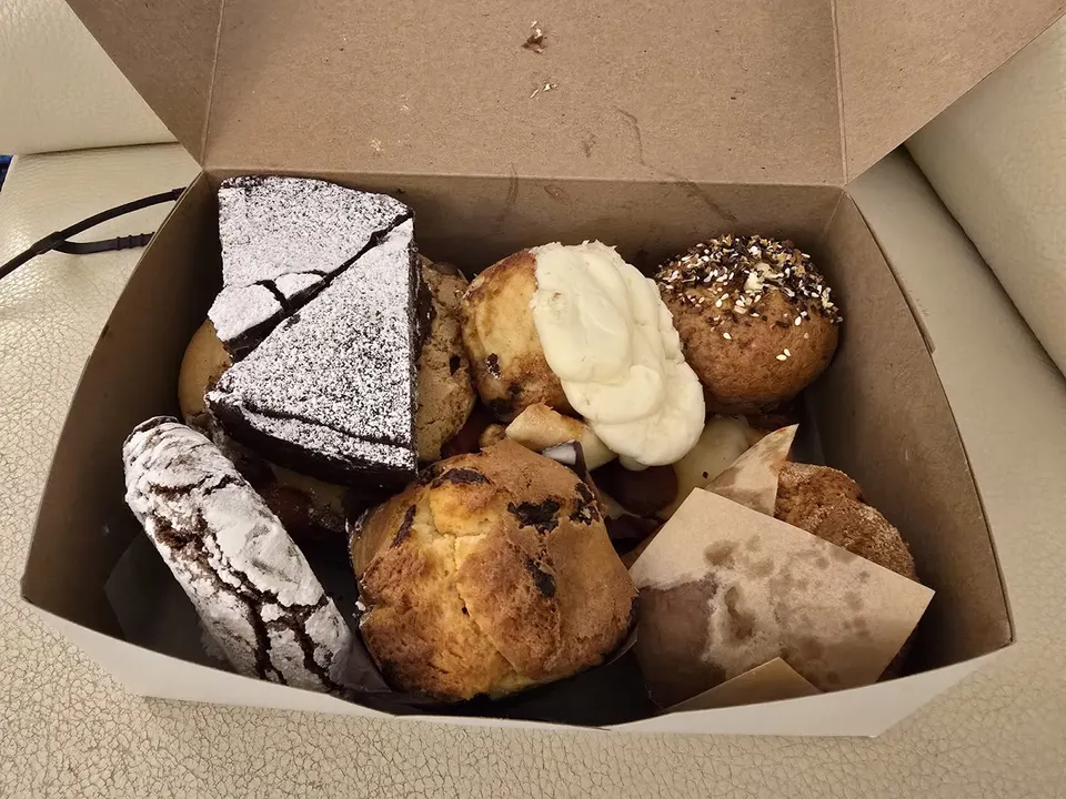 A cardboard bakery box filled with an assortment of baked goods, including powdered sugar-dusted chocolate cake slices, muffins, and cookies.