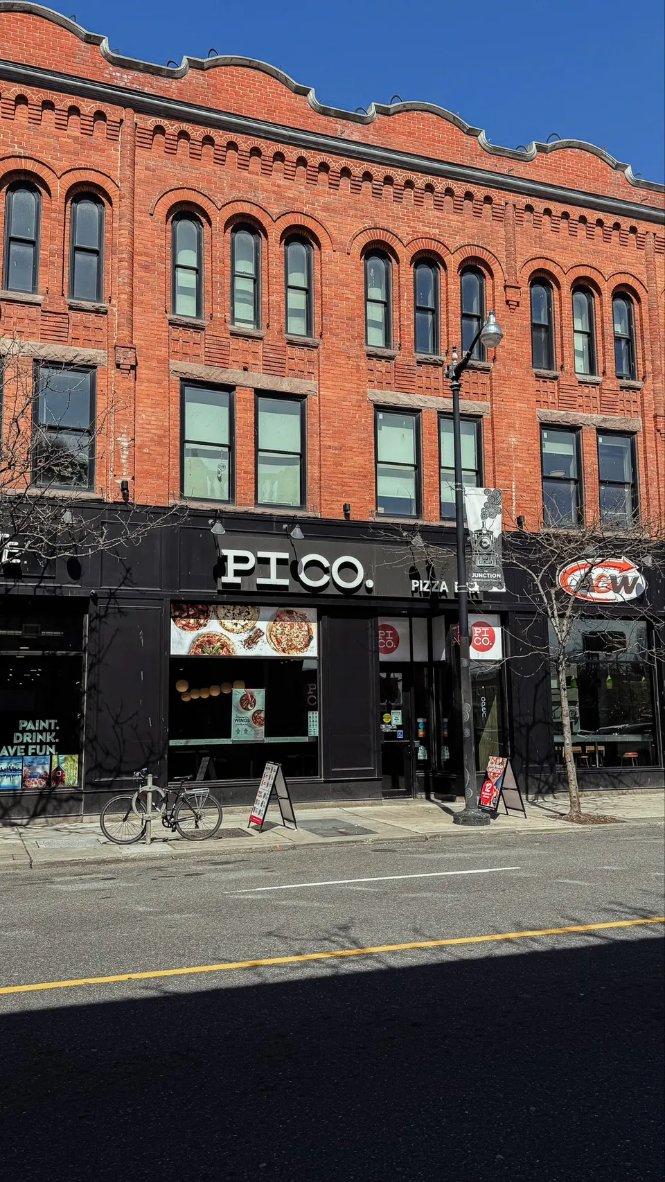 Exterior of Pi Co. Pizza Bar, a restaurant with a black storefront and large white lettering, located on the ground floor of a historic red brick building with arched windows.