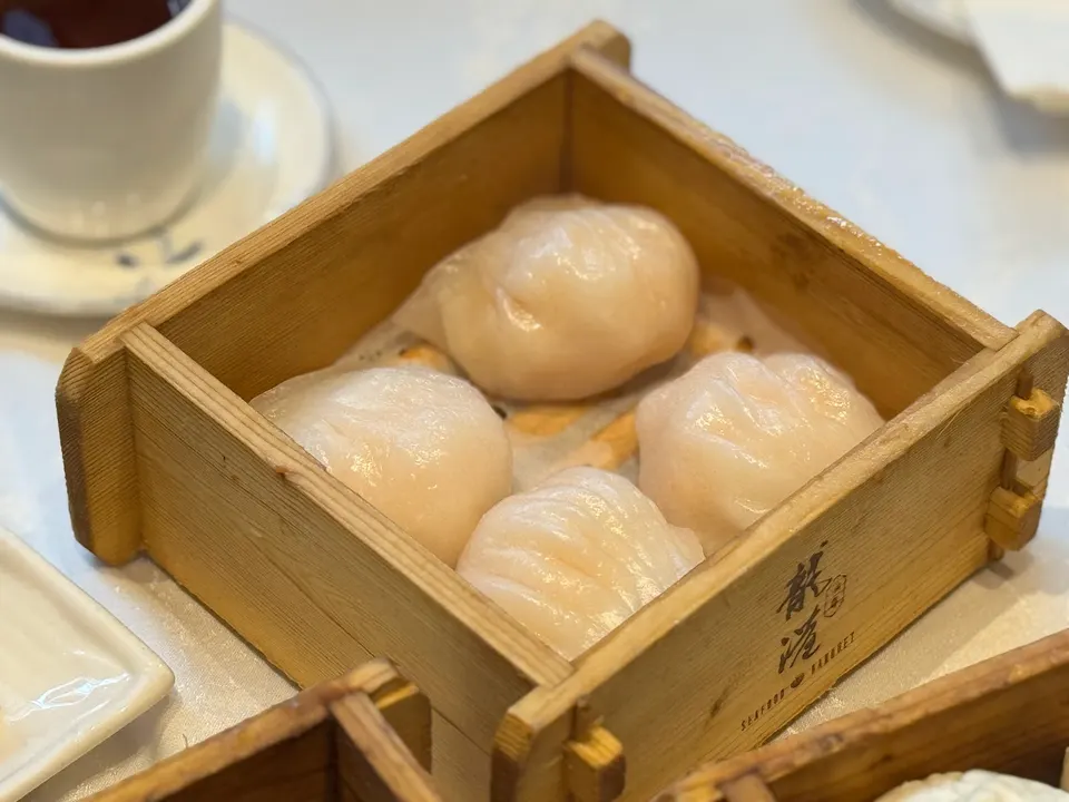 Four translucent shrimp dumplings, or har gow, served hot inside a square wooden steamer basket on a restaurant table.