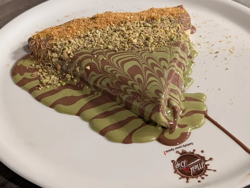 A triangular crepe on a white plate, covered in a marbled pattern of chocolate and pistachio sauce, topped with crushed pistachios and shredded pastry.