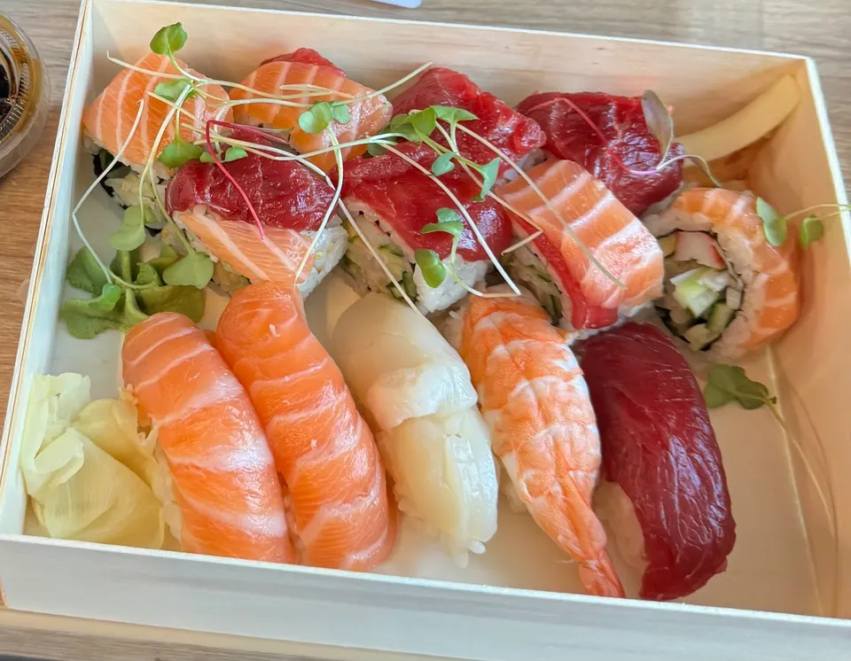 An assortment of sushi in a light wooden box, featuring salmon, tuna, and shrimp nigiri alongside various maki rolls topped with fresh fish and microgreens.