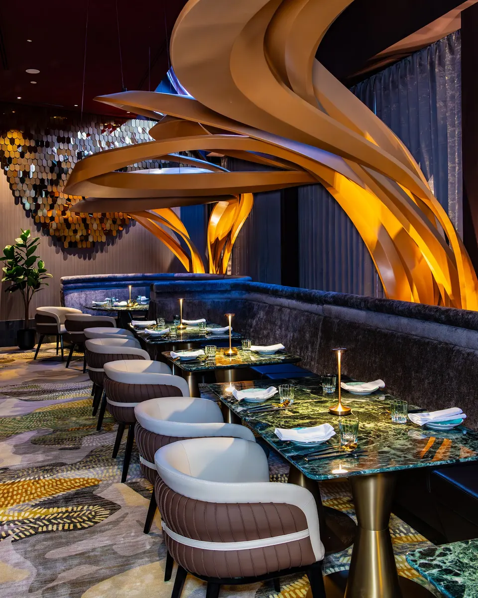 An upscale dining area featuring green marble tables, plush curved booths, and modern armchairs under a dramatic, flowing golden ceiling sculpture.