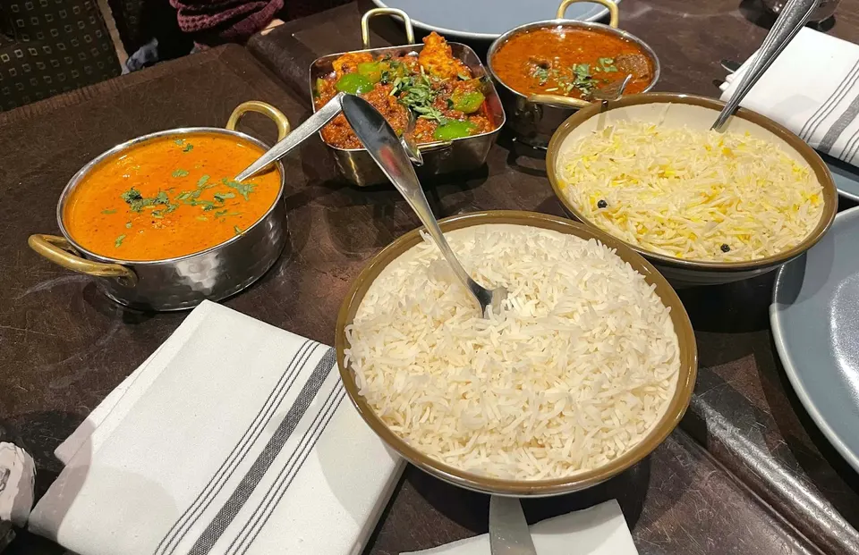 A spread of Indian dishes on a dark table, including bowls of white basmati rice, yellow saffron rice, butter chicken, and other curries with serving spoons.