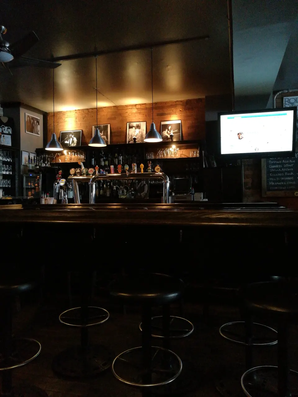 A dimly lit bar counter with black leather barstools, silver beer taps, and three hanging pendant lights illuminating framed black-and-white photos on a brick wall.