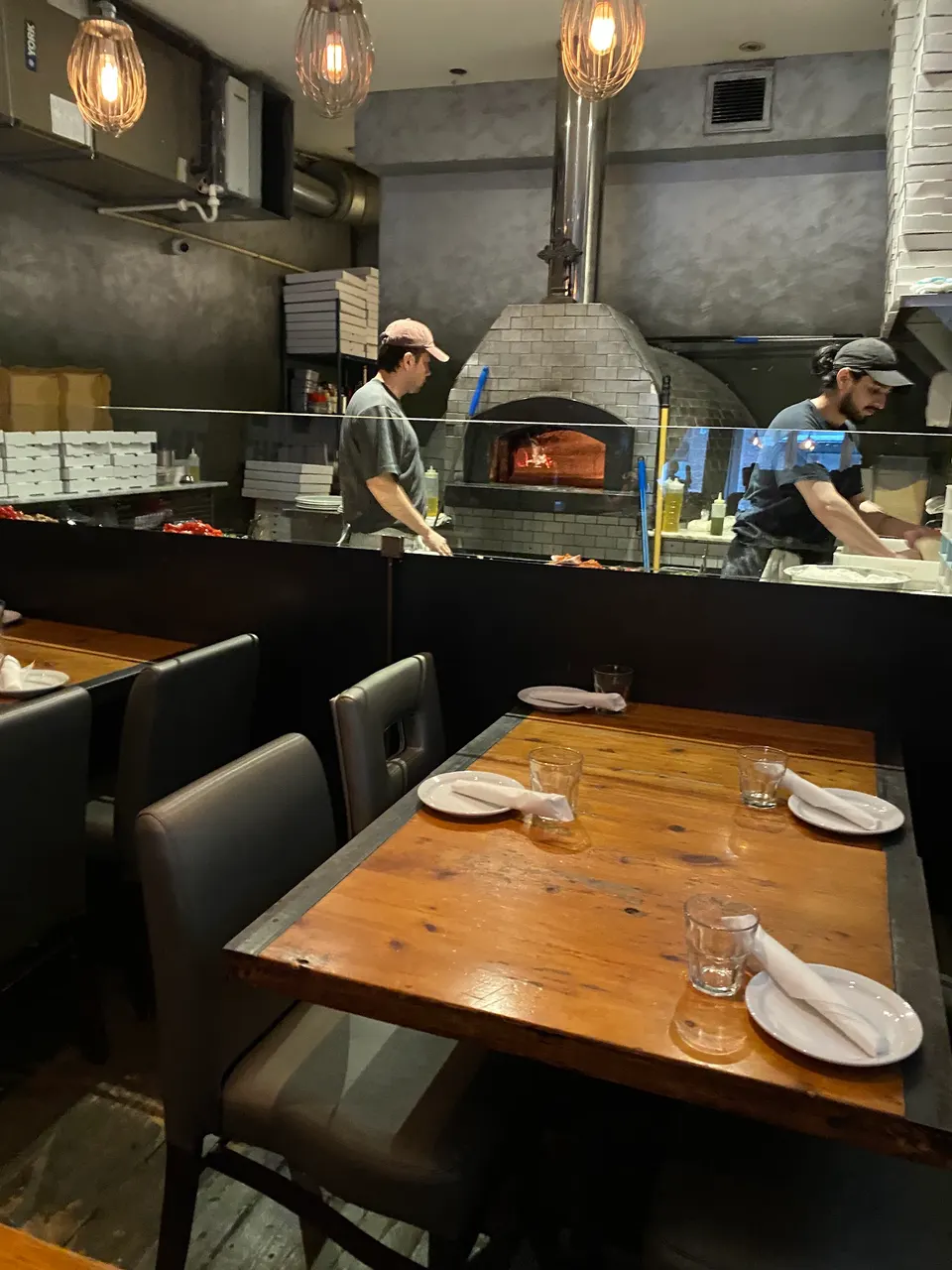 A dining area at Pizzeria Defina featuring a wooden table set with plates and glasses, with a view of the kitchen where staff are preparing food near a brick pizza oven.