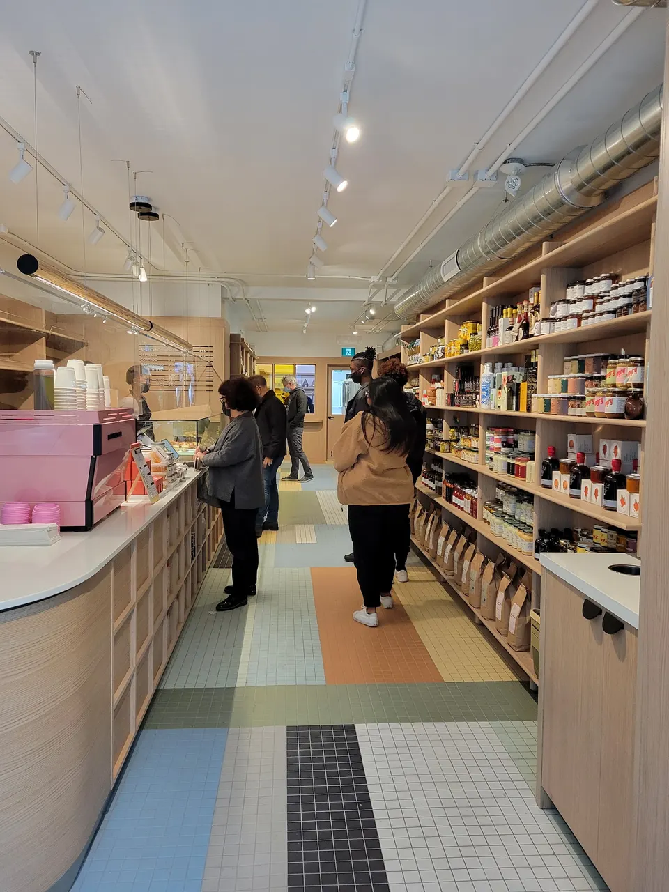 Interior view of Dear Grain bakery showing customers waiting in line along a wooden counter, with shelves stocked with artisanal goods on the right and a colorful tiled floor.