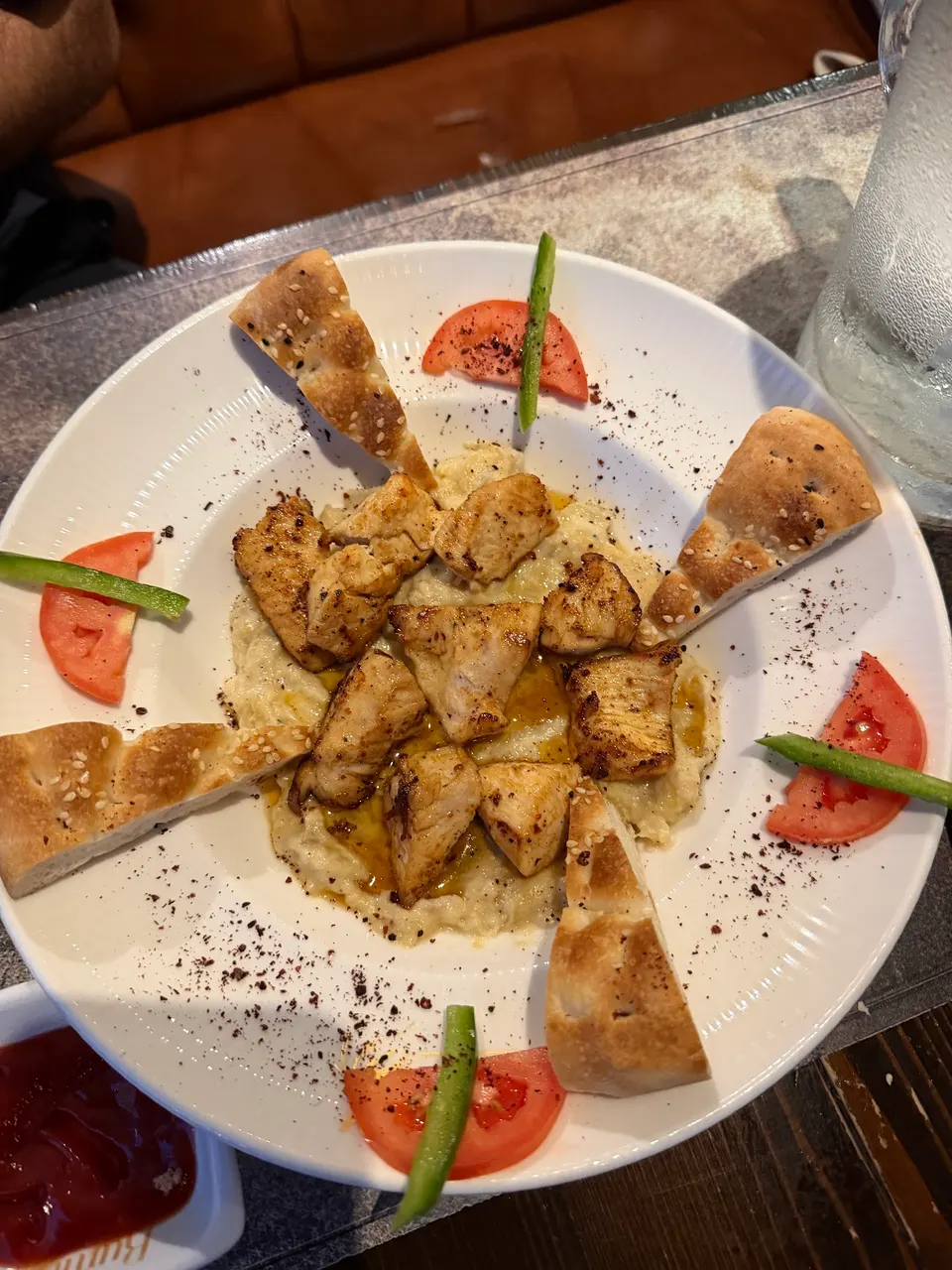 A white plate featuring grilled chicken pieces served over a creamy dip, garnished with wedges of flatbread, tomato slices, and green pepper strips arranged in a star pattern.