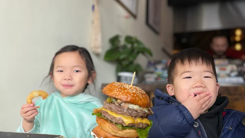 Two young children sitting at a restaurant table, one holding an onion ring and the other smiling with a hand to their mouth, next to a very tall double cheeseburger stacked with onion rings.