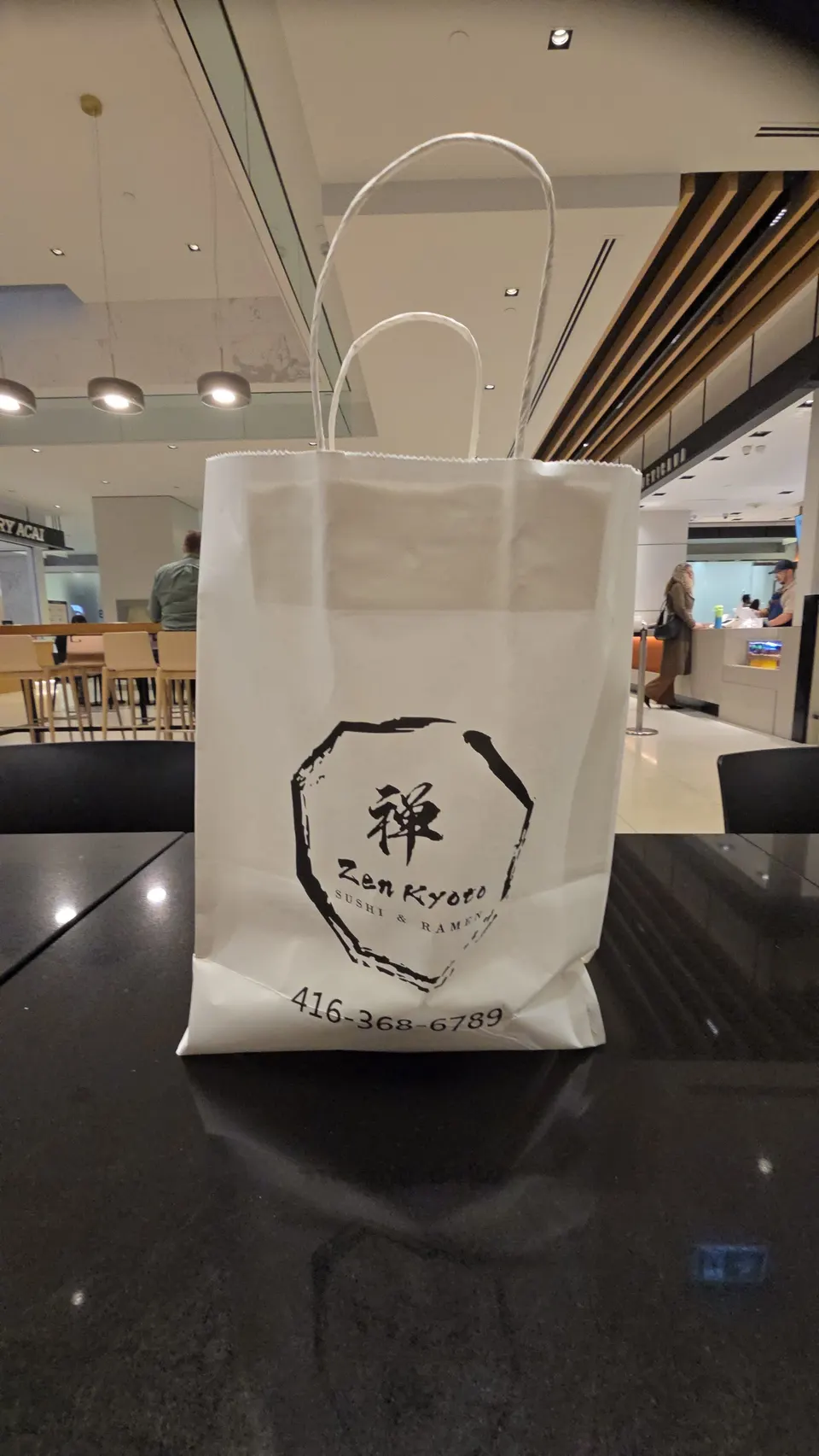 A white paper takeout bag from Zen Kyoto Sushi & Ramen sitting on a dark, reflective table in a food court setting.