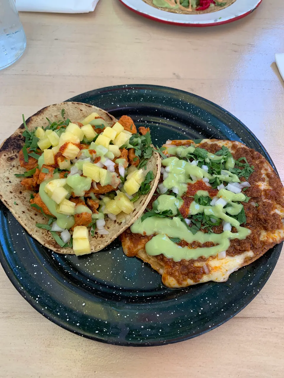 Two tacos served on a dark green speckled plate. One taco features seasoned chicken topped with diced pineapple, onions, cilantro, and green salsa. The other is a flat tostada-style taco with seasoned ground meat, melted cheese, onions, cilantro, and a drizzle of green salsa.