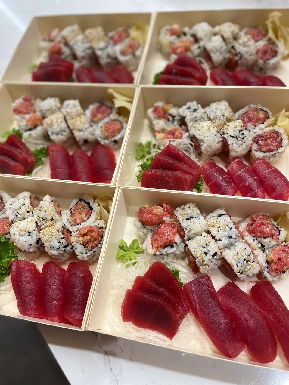 Six wooden bento boxes arranged in a grid, each filled with fresh tuna sashimi slices, spicy tuna maki rolls, and sesame-sprinkled sushi rolls on a bed of shredded daikon radish.