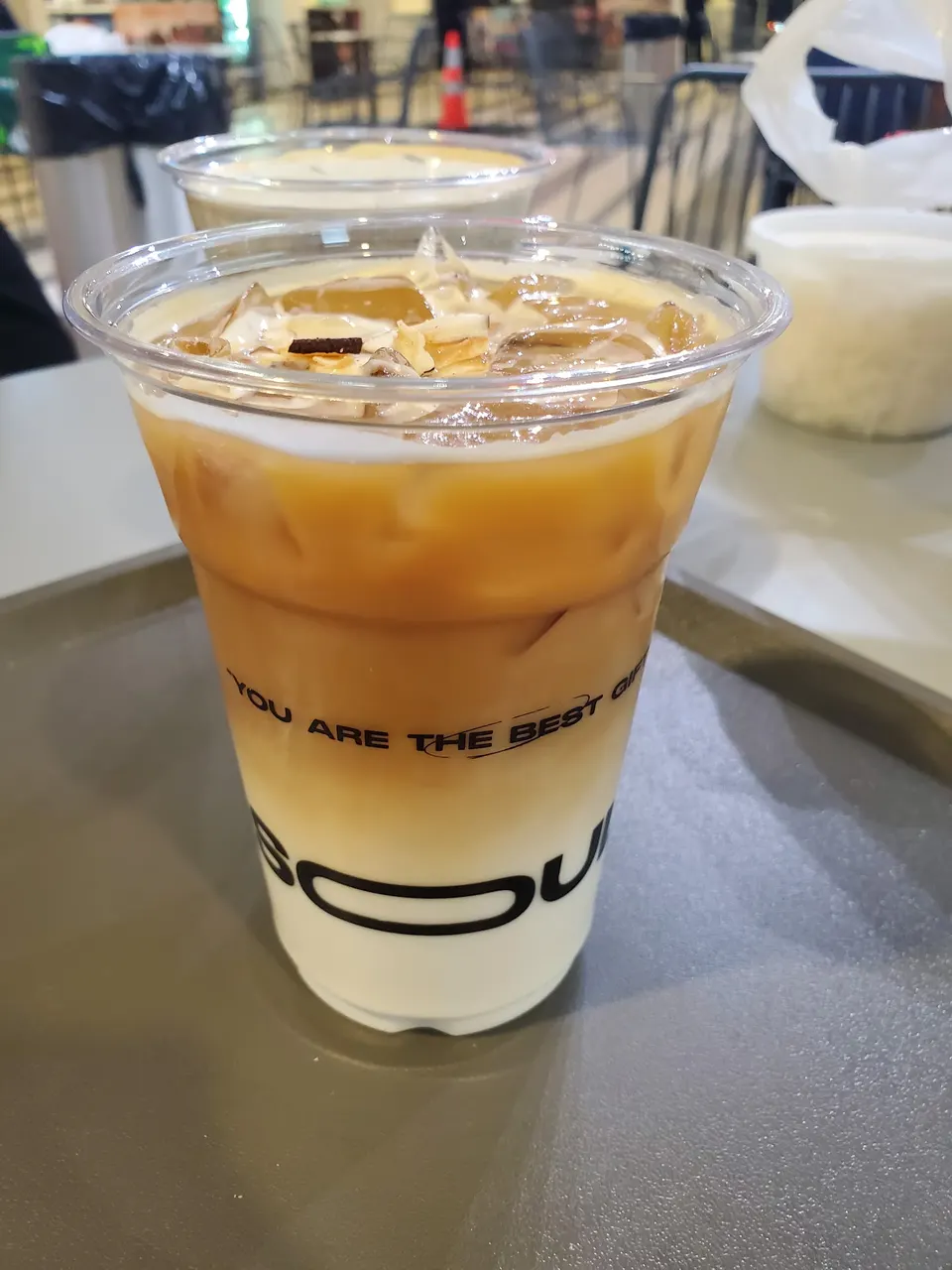 An iced latte in a clear plastic cup with distinct layers of white milk at the bottom and light brown espresso on top, garnished with sliced almonds and ice cubes.