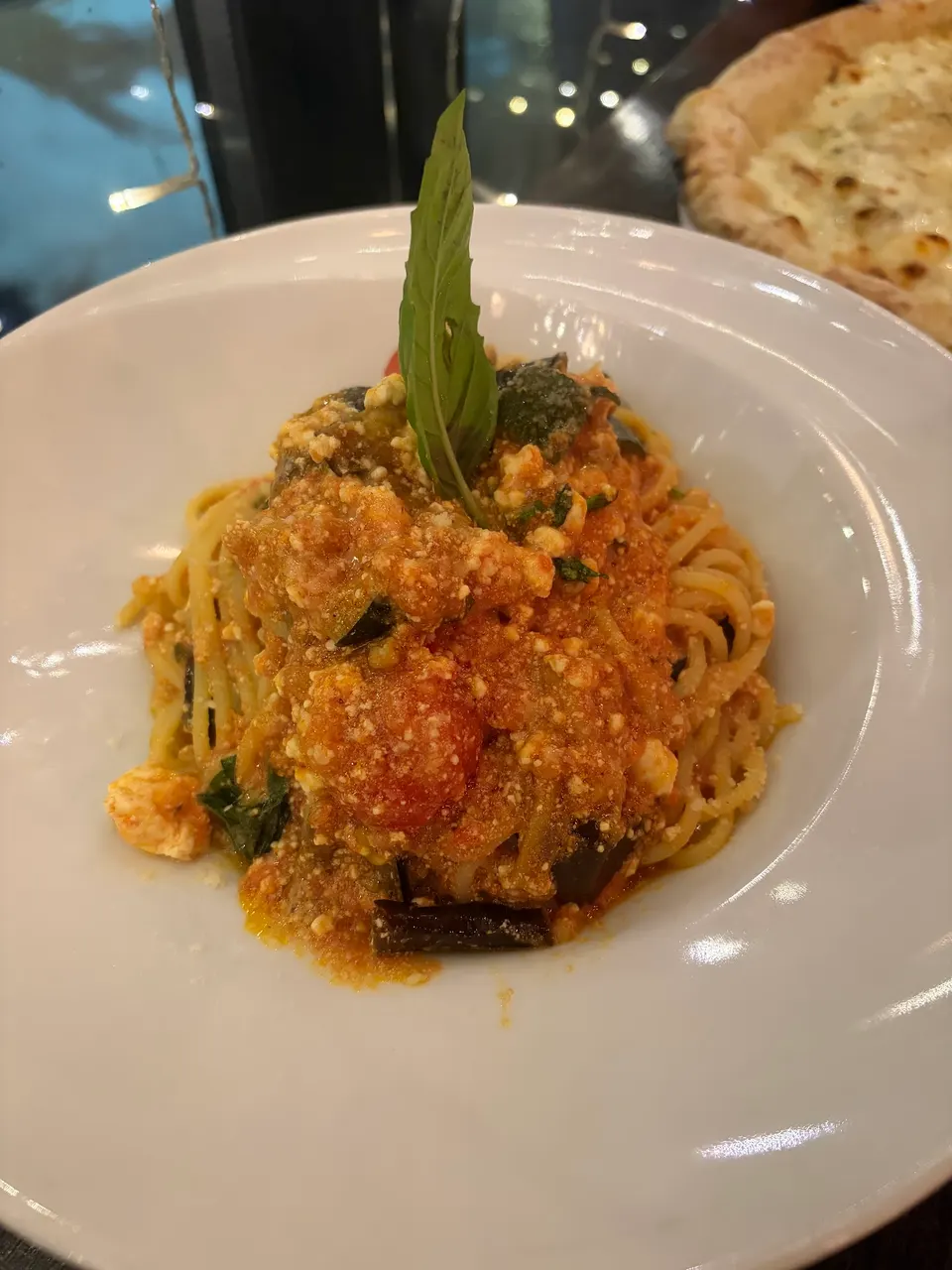 A plate of spaghetti pasta tossed in a rich tomato sauce with melted cheese, eggplant, and fresh basil leaves, served in a white bowl at SIP Wine Bar Restaurant.