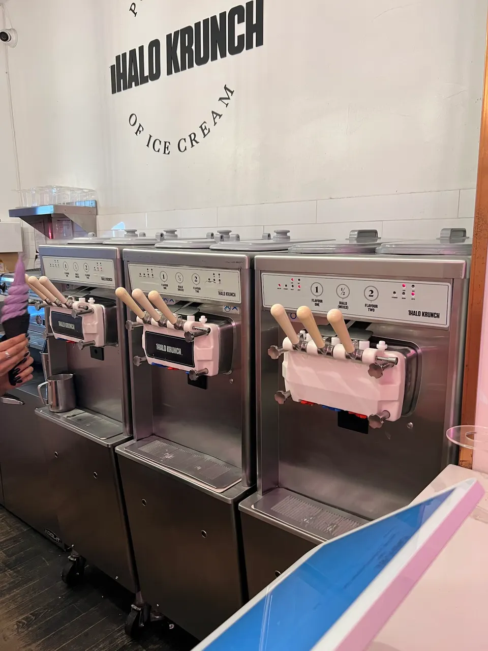 Three stainless steel soft-serve ice cream machines lined up against a white wall with the iHalo Krunch logo, with a hand holding a purple soft-serve cone on the left.