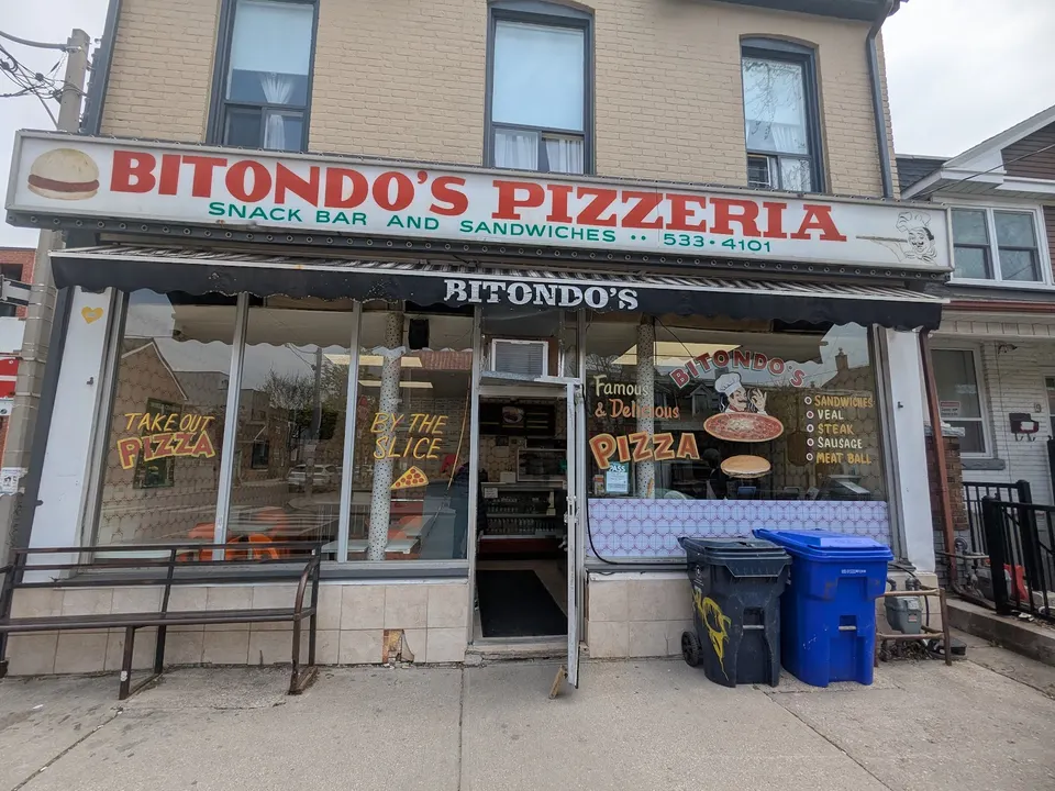 Street-level exterior view of Bitondo's Pizzeria, featuring a large white sign with red lettering, a black awning, and window decals advertising pizza by the slice and sandwiches.