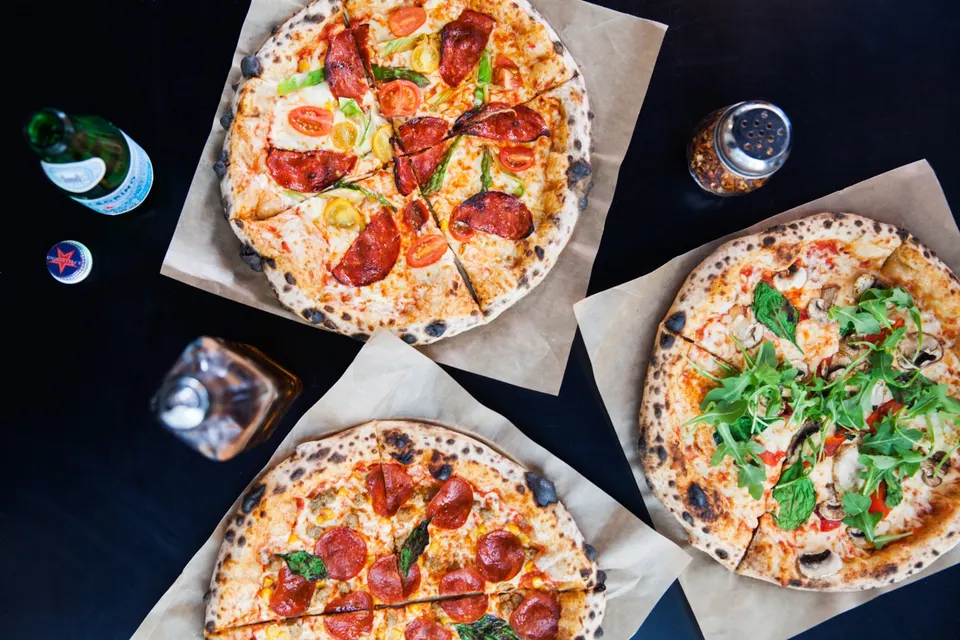 An overhead view of three freshly baked thin-crust pizzas on brown parchment paper, served on a dark table alongside a bottle of sparkling water, a bottle of oil, and a red pepper shaker.
