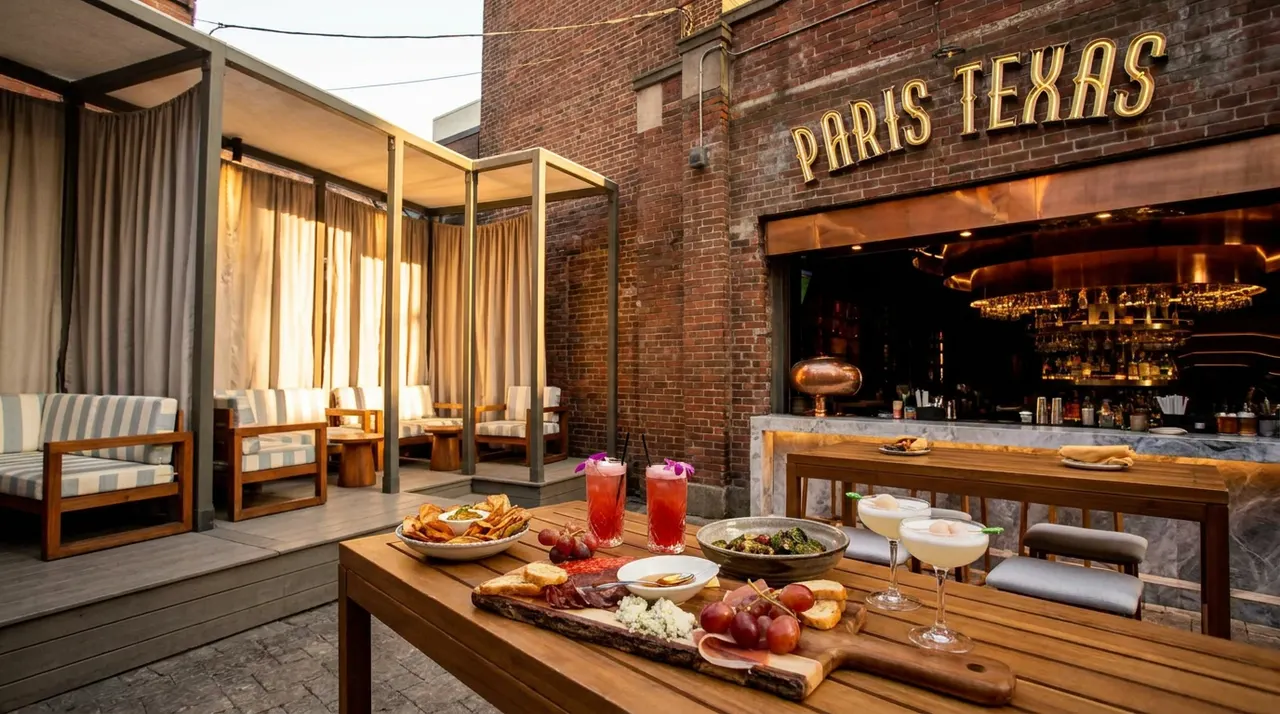 An outdoor patio at Paris Texas with wooden tables set with charcuterie, cocktails, and appetizers, featuring a brick wall with a neon sign and covered cabana seating in the background.