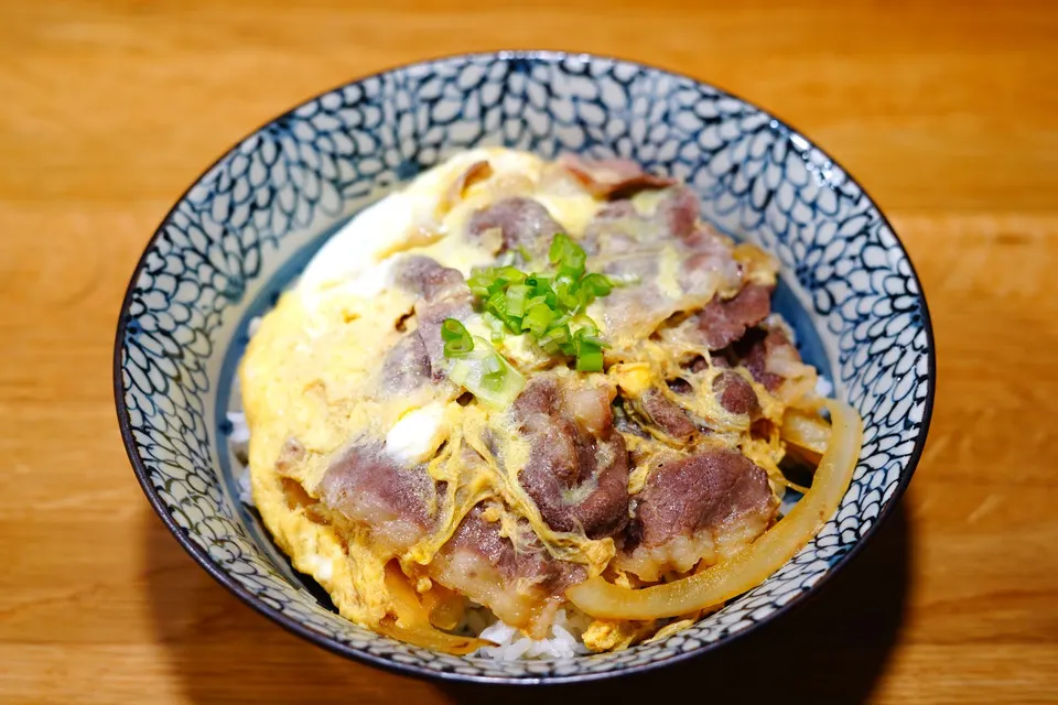 A Japanese beef and egg rice bowl, gyudon, served in a blue patterned ceramic bowl on a wooden table, garnished with chopped green onions.
