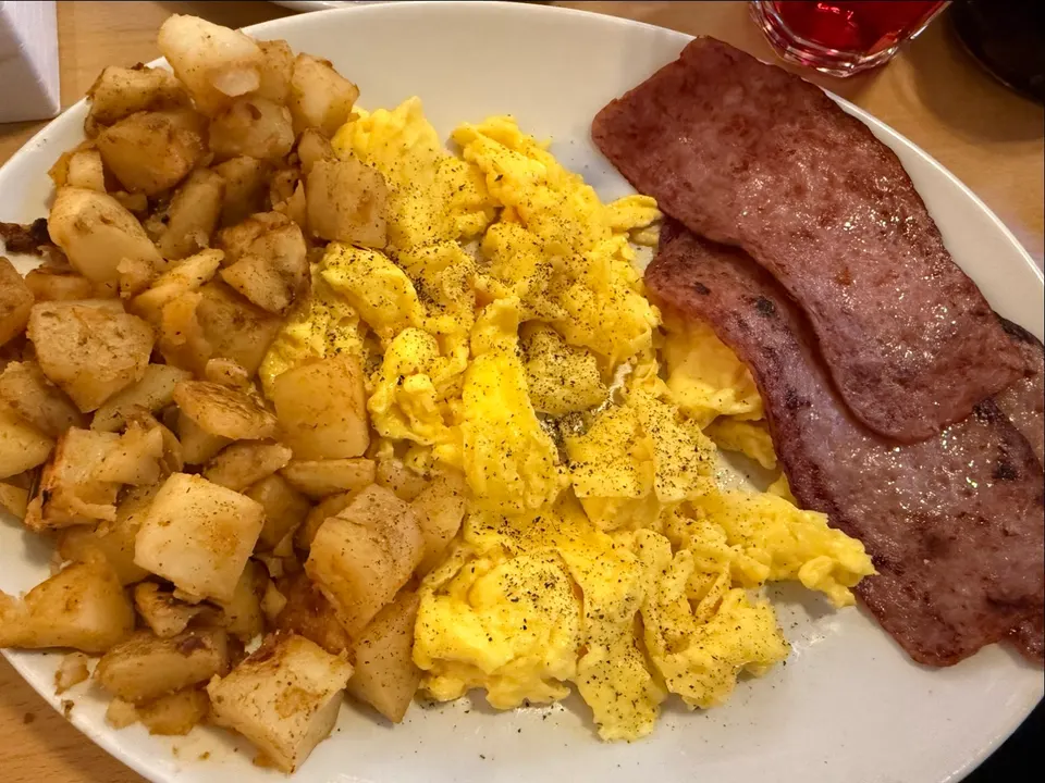 A white plate filled with breakfast food including seasoned home fries on the left, scrambled eggs with black pepper in the center, and strips of turkey bacon on the right.