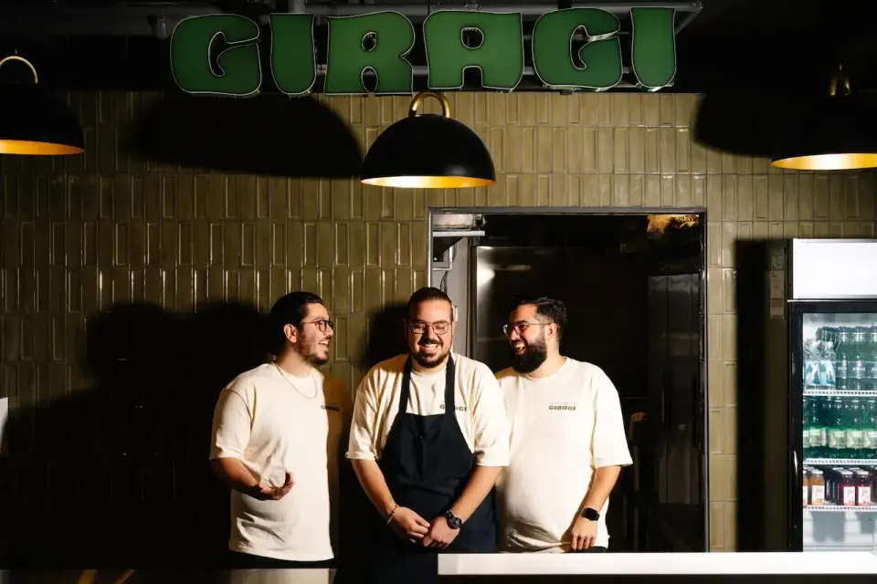 Three smiling men, one in an apron, stand behind a counter under a green 'GIRAGI' sign, with olive green tiled walls and pendant lights in the background.