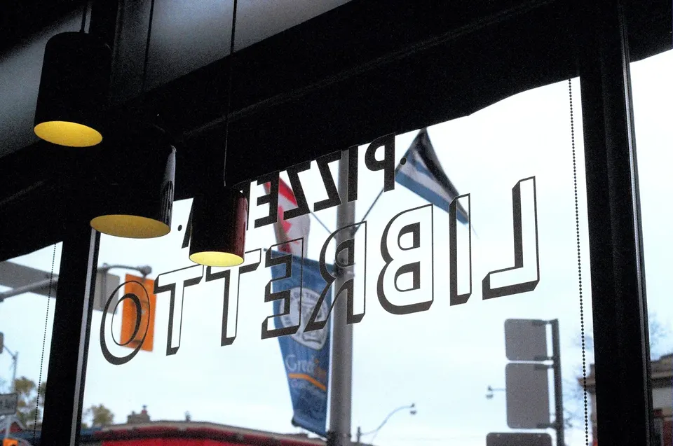 A view looking out a window of Pizzeria Libretto, showing the restaurant's name printed in reverse on the glass, with hanging pendant lights in the foreground and street elements outside.