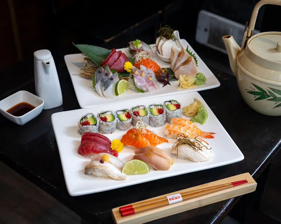 Two white rectangular plates filled with an assortment of fresh sushi, sashimi, and maki rolls, served alongside a small bowl of soy sauce, a teapot, and chopsticks on a dark table.