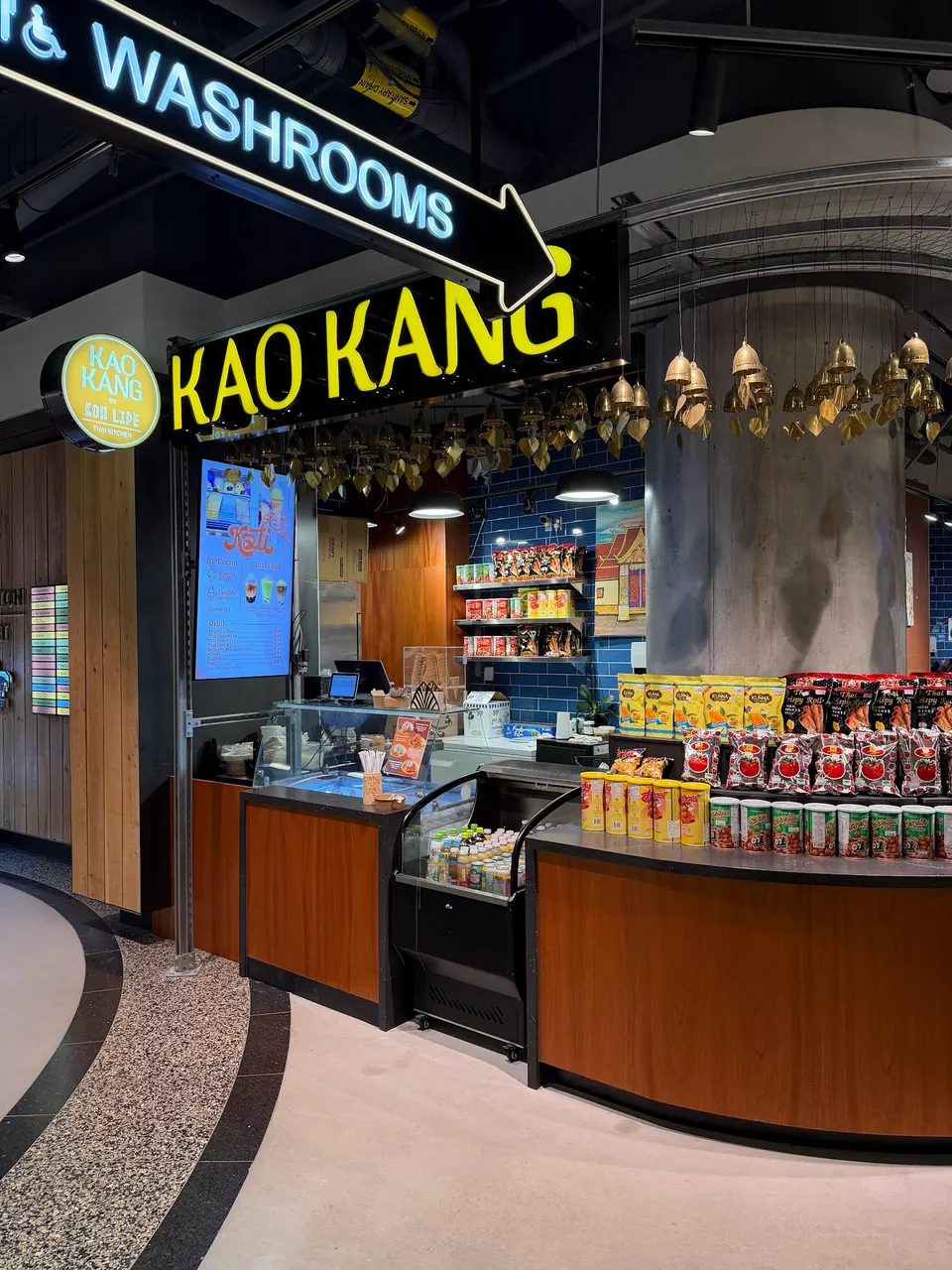 The storefront of Kao Kang by Koh Lipe, featuring a bright yellow sign, a display counter with Thai snacks and drinks, and a blue tiled wall in the background.