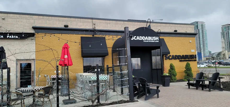Exterior view of Scaddabush Italian Kitchen & Bar in Scarborough, featuring a yellow brick facade, black awnings, outdoor patio seating with tables and chairs, and a black entryway canopy.