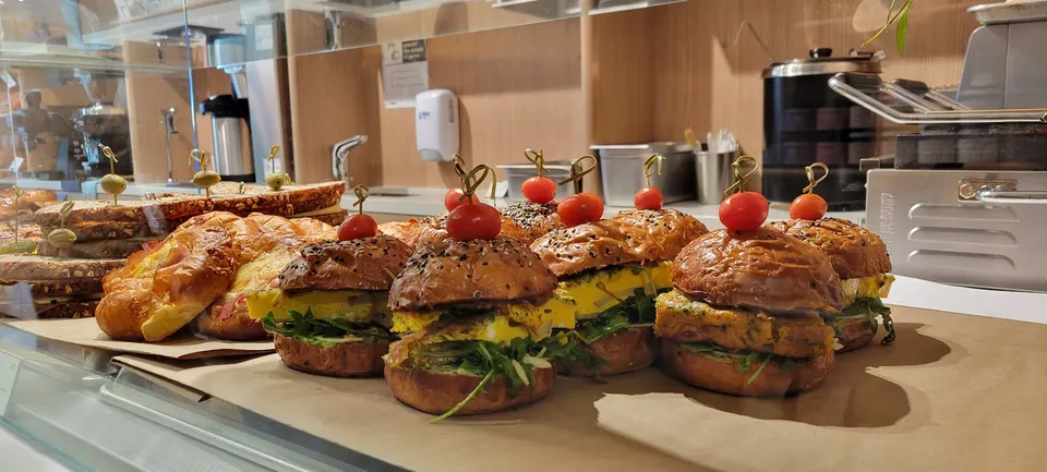 A row of small breakfast sandwiches on sesame buns with egg frittata, greens, and cherry tomatoes on toothpicks, displayed on brown paper in a bakery display case.