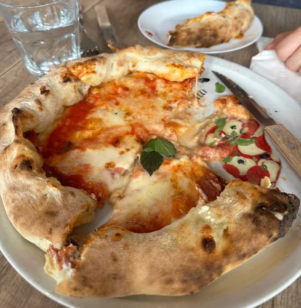 A freshly baked Neapolitan-style pizza with a thick, puffed crust, melted mozzarella cheese, tomato sauce, and a fresh basil leaf, served on a white plate with a knife beside it.
