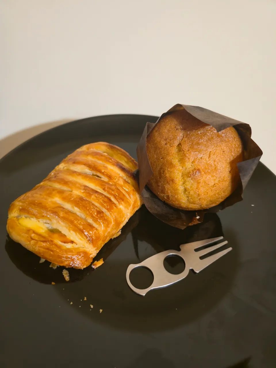 A golden-brown puff pastry with a lattice top next to a muffin in a dark paper liner, served on a black plate with a small metal fork.