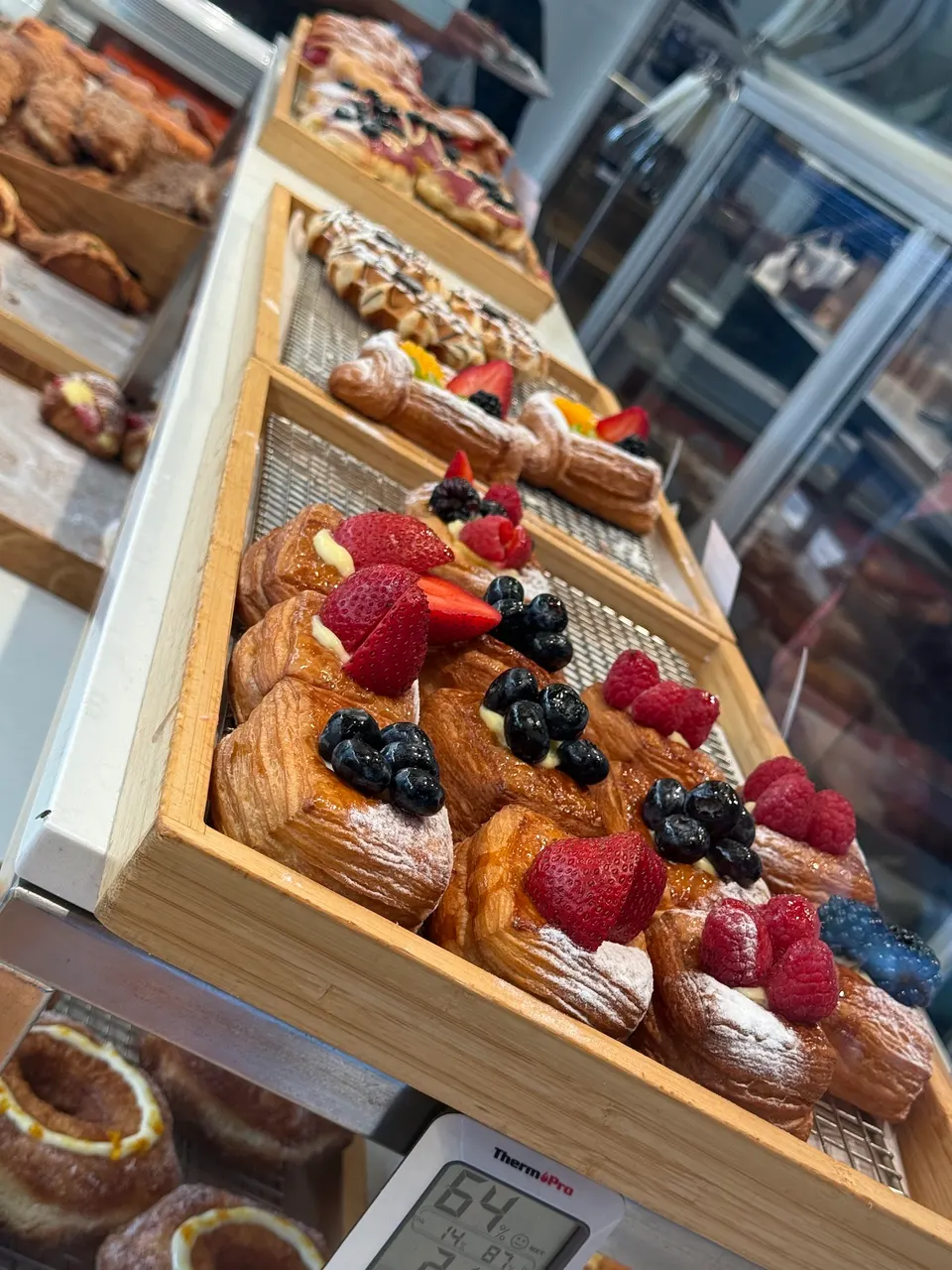 A close-up, angled shot of a wooden bakery display tray filled with fresh fruit pastries topped with strawberries, blueberries, and raspberries, with other pastries visible in the background.