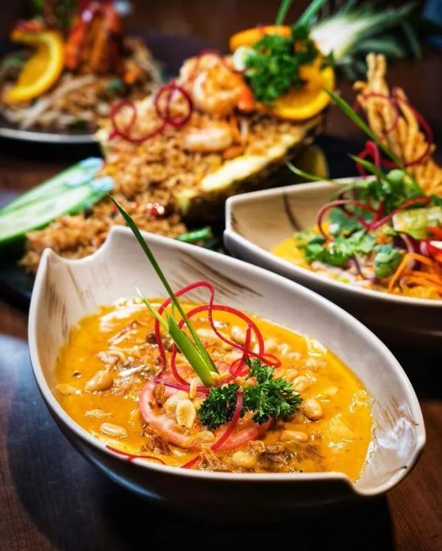 A close-up of a bowl of yellow Thai curry garnished with peanuts, fresh herbs, and red chili curls, with pineapple fried rice and other Thai dishes blurred in the background.