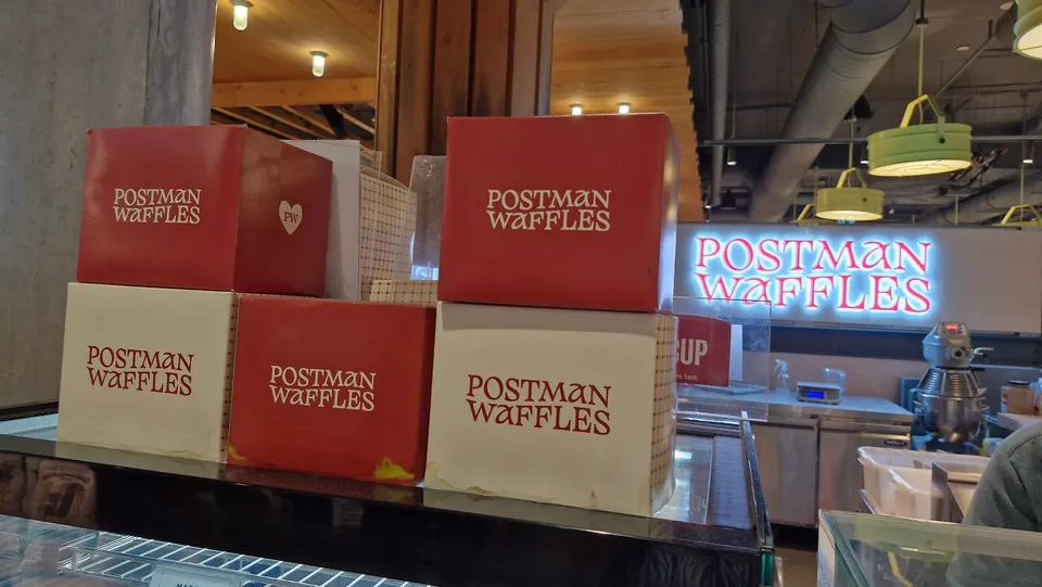 Several red and white boxes with the 'Postman Waffles' logo stacked on a counter, with a neon sign for 'Postman Waffles' glowing in the background. A kitchen area with stainless steel equipment is visible behind the counter.