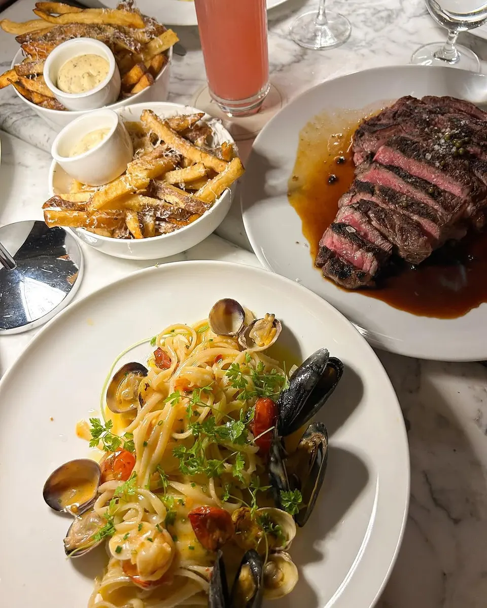 A marble table set with a plate of seafood linguine with clams and mussels, a sliced steak with rich gravy, two bowls of truffle fries with dipping sauces, and a pink cocktail.