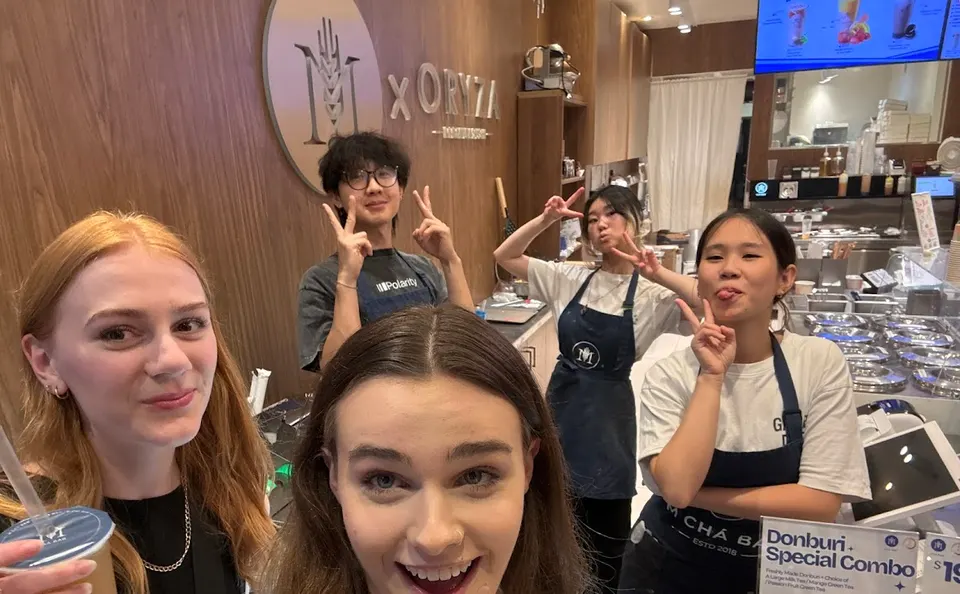 A group of five young people, including staff in aprons, smiling and making peace signs for a selfie inside ORYZA Sushi Bar.