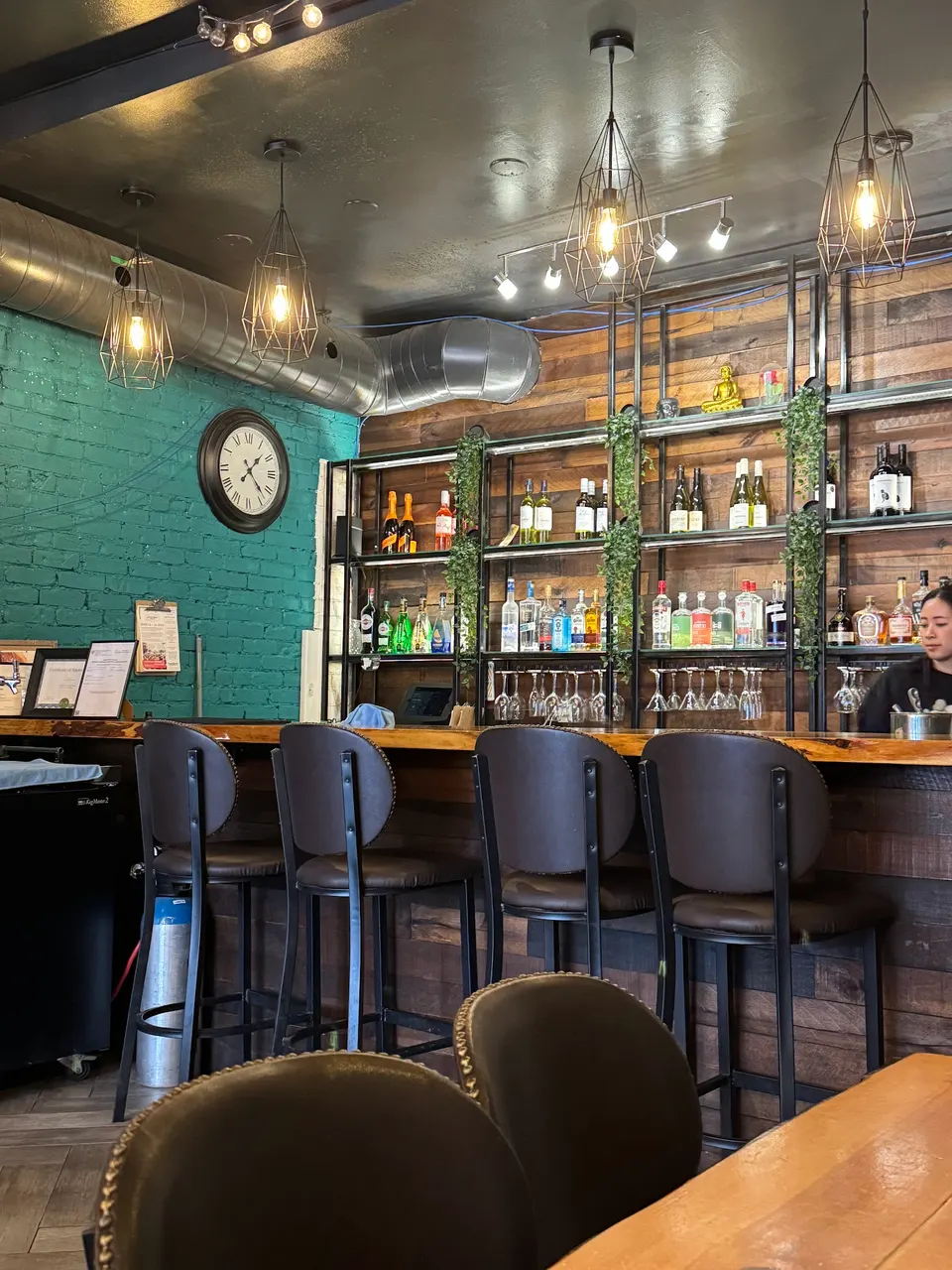 A cozy bar area at Imm Thai Kitchen featuring dark leather bar stools, a wooden bar counter, shelves stocked with liquor bottles against a wood-paneled wall, a green brick accent wall with a clock, and industrial pendant lights.