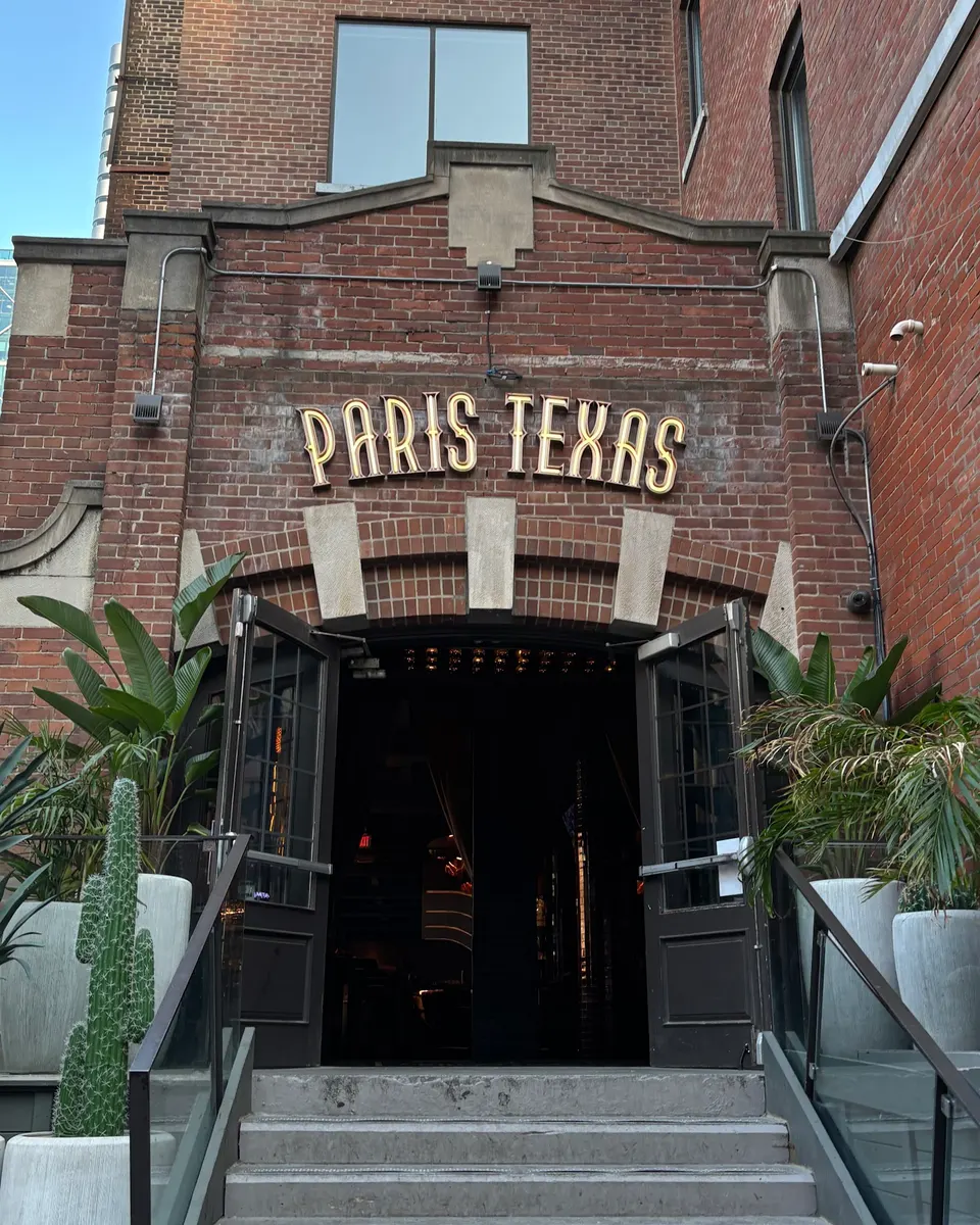The brick exterior entrance of Paris Texas bar in Wellington Place, featuring a glowing neon sign above double doors, flanked by potted tropical plants and a cactus.