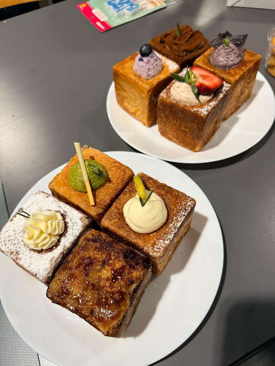 Two white plates on a grey table, each holding four square-shaped cube croissants with various toppings like cream, fresh strawberry, blueberry, and caramelized sugar.